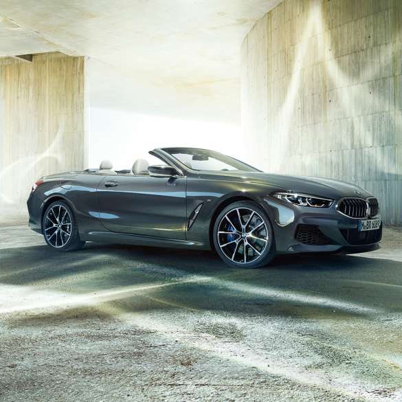 BMW M850i xDrive Cabrio, vista de tres cuartos del lateral F91. BMW M850i xDrive Cabrio en Dravitgrau metalizado con la capota abierta, vista de tres cuartos del lateral, parado delante de un edificio de hormigón F91.