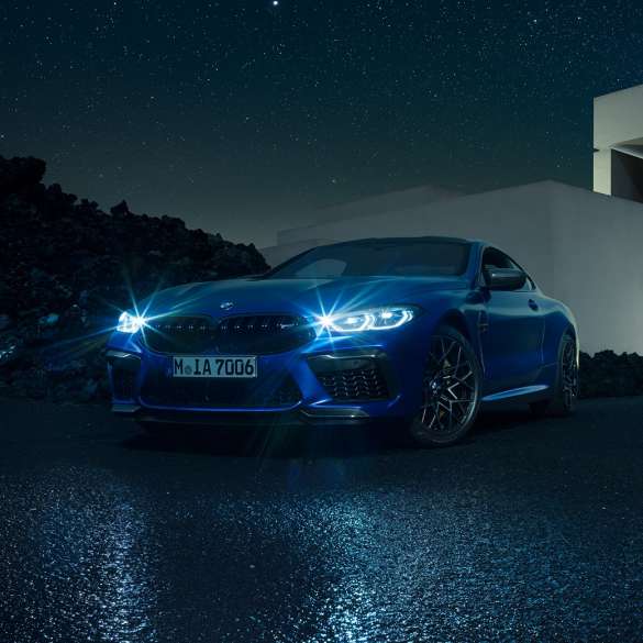 BMW M8 Competition Coupé, vista de tres cuartos del frontal. BMW M8 Competition Coupé en Marina Bay Blau metalizado con los faros LED encendidos, vista de tres cuartos del frontal, parado delante de una casa de campo, de noche.