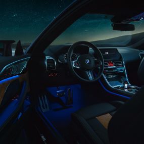 Interior del BMW M8 Competition Coupé. BMW M8 Competition Coupé, interior de noche, volante M de cuero y luz ambiente azul con la puerta del conductor abierta.