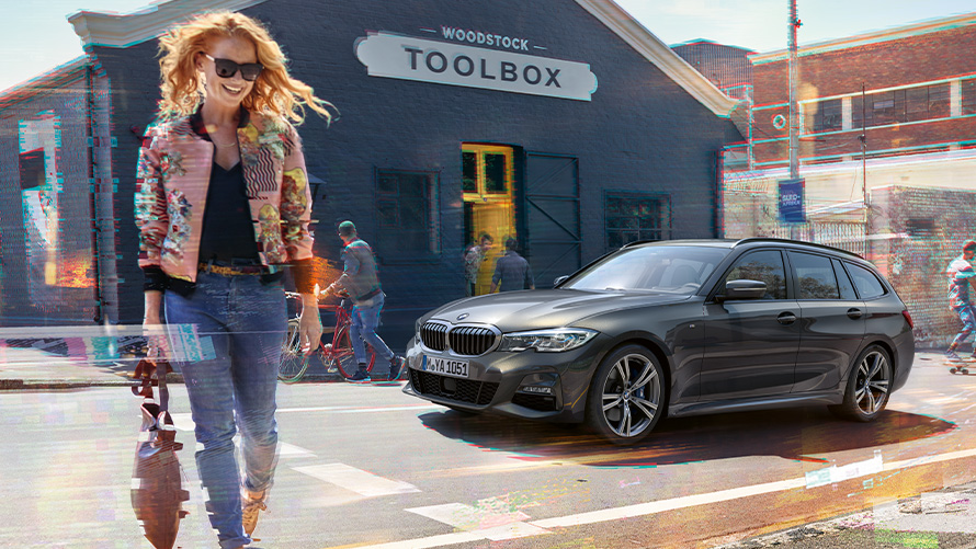 BMW Serie 3 Touring (G21), vista de tres cuartos del frontal en la calle con mujer delante de un establecimiento de herramientas de madera Mujer cruza la calle frente a un BMW Serie 3 Touring (G21) y un establecimiento de herramientas de madera.