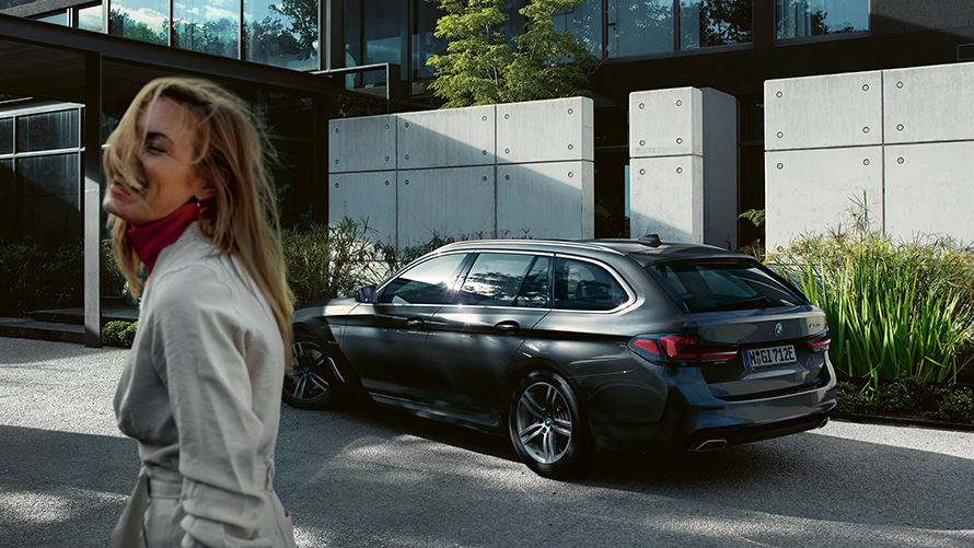 Mujeres BMW Serie 5 Touring G31 LCI Sophisto Grey efecto brillante metallic, vista lateral 7/8 Mujer delante de un BMW Serie 5 Touring aparcado G31 Renovación 2020 en Sophisto Grey efecto brillante metallic, vista lateral 7/8