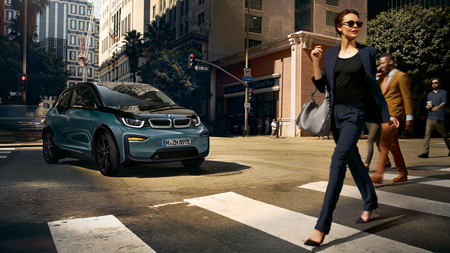 BMW i3 I01 Vista de tres cuartos del frontal, con mujer cruzando por un paso de cebra BMW i3 I01 Blue Ridge Mountain metallic con detalles Frozen Grey metallic, vista de tres cuartos del frontal, con mujer cruzando por un paso de cebra