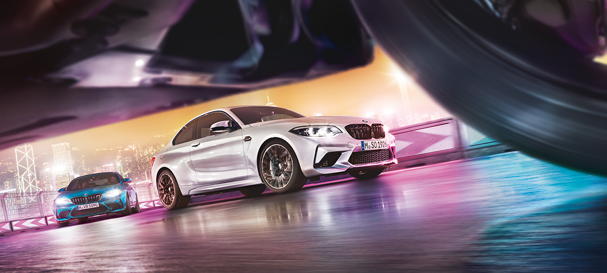 El BMW M2 Competition sobre el asfalto reflectante. Dos vehículos BMW M2 Competition derrapando en el circuito, con el perfil nocturno de la ciudad de fondo, en una vista desde el suelo.