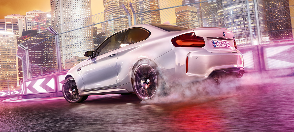 El BMW M2 Competition Coupé acelera BMW M2 Competition en blanco derrapando en el circuito con el perfil nocturno de la ciudad de fondo.