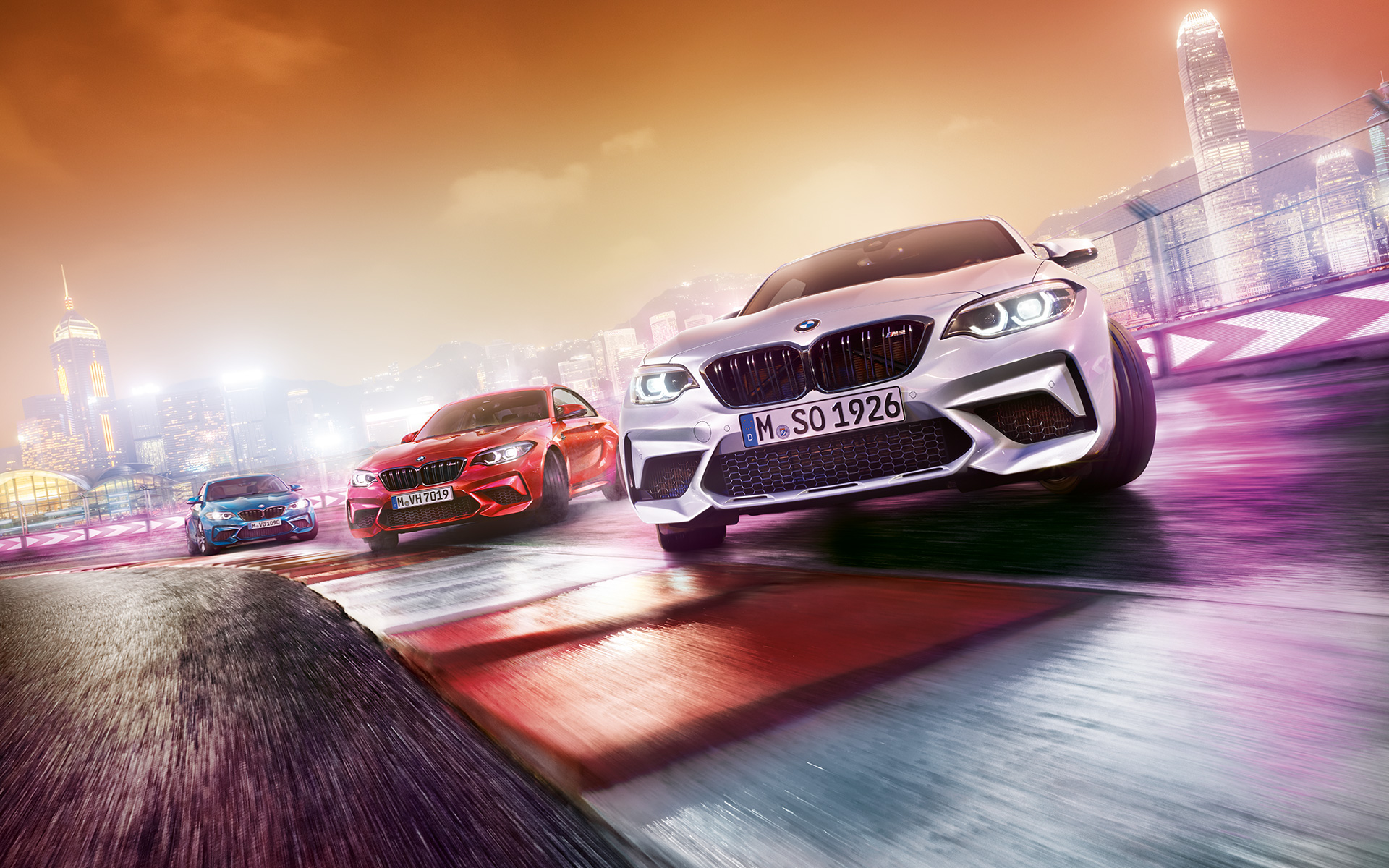 El BMW M2 Competition en acción Tres vehículos BMW M2 Competition derrapando en el circuito con el perfil nocturno de la ciudad de fondo.