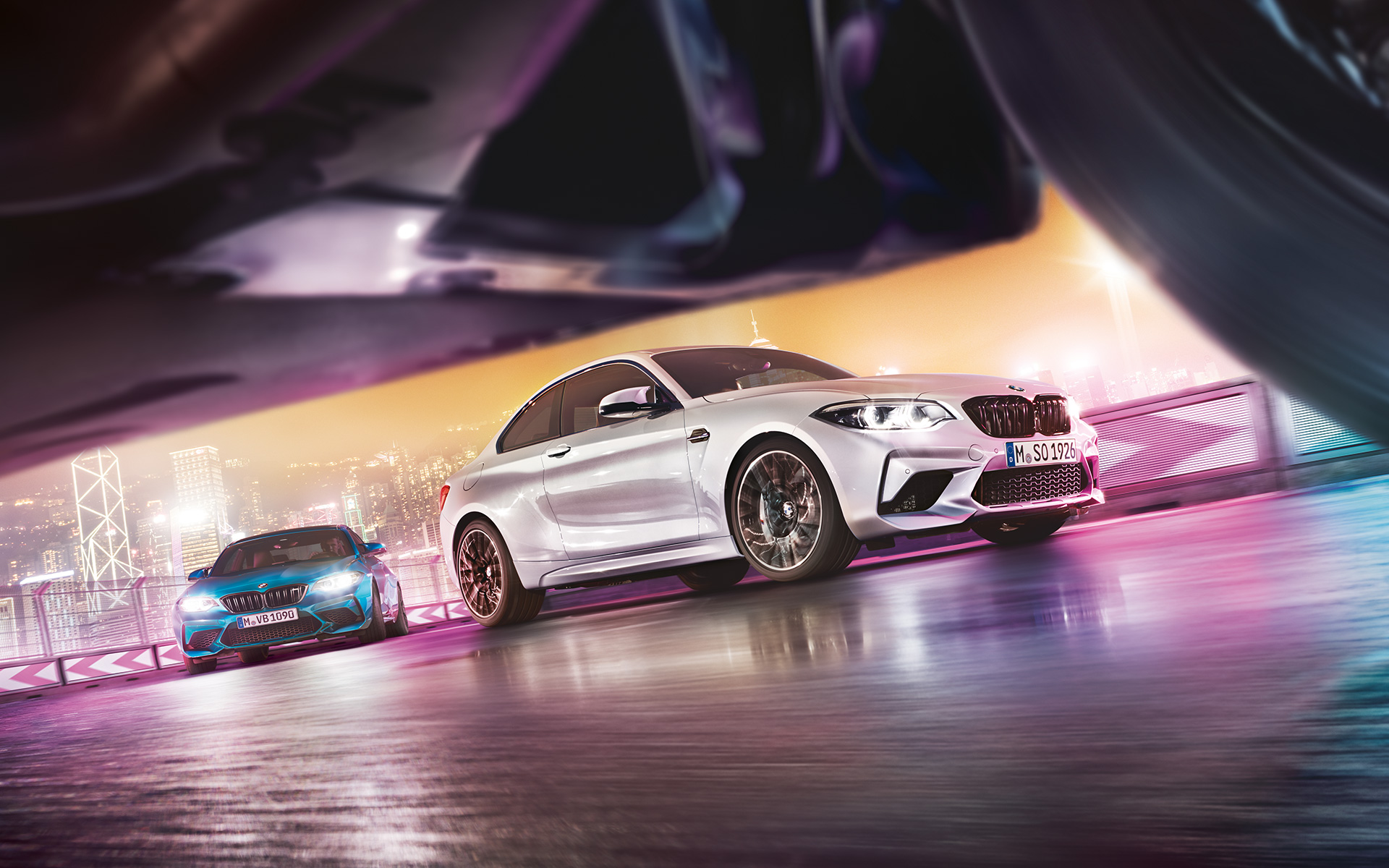 El BMW M2 Competition sobre el asfalto reflectante. Dos vehículos BMW M2 Competition derrapando en el circuito, con el perfil nocturno de la ciudad de fondo, en una vista desde el suelo.