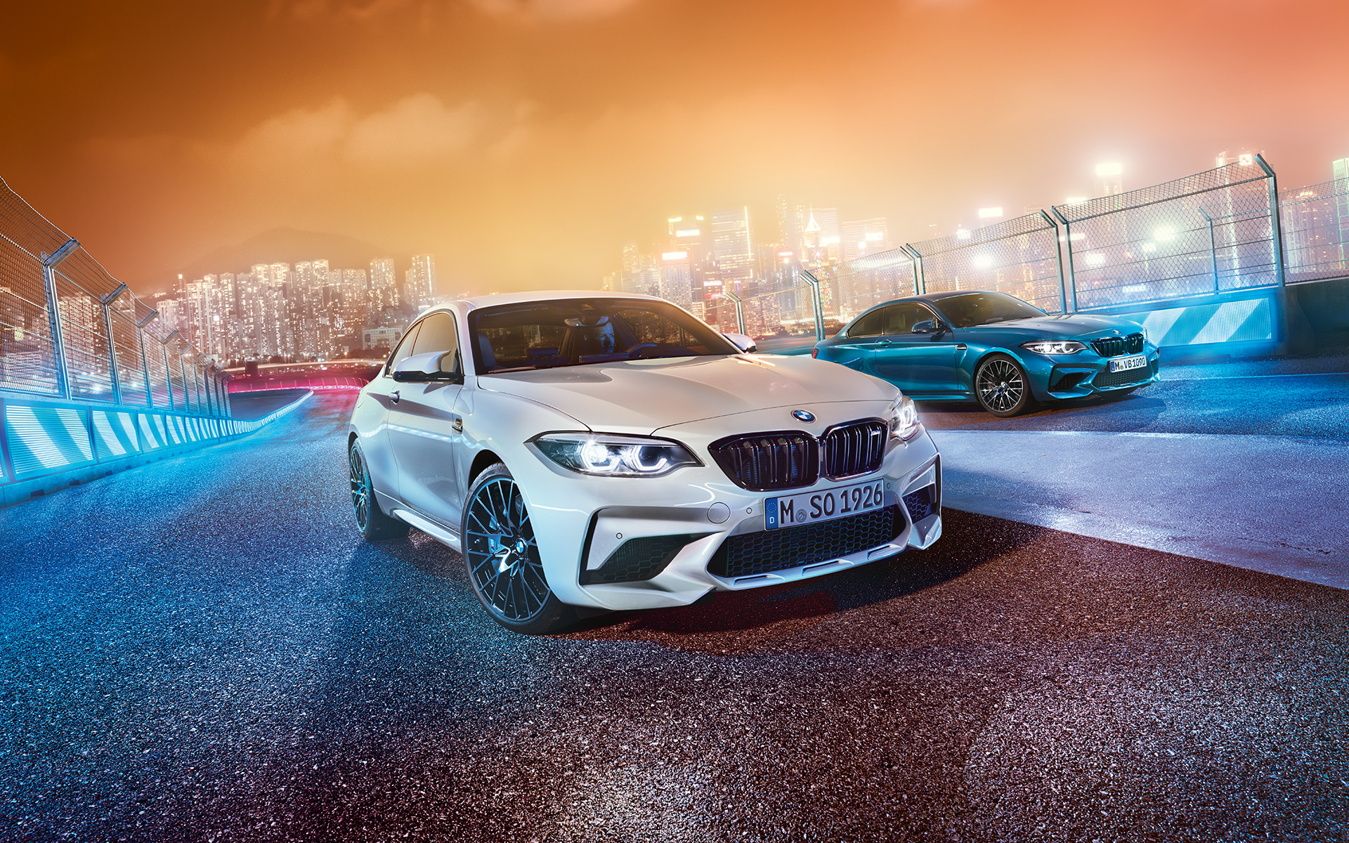 Dos vehículos BMW M2 Competition en el circuito BMW M2 Competition en blanco y azul, con el perfil nocturno de la ciudad de fondo, en una vista delantera oblicua.