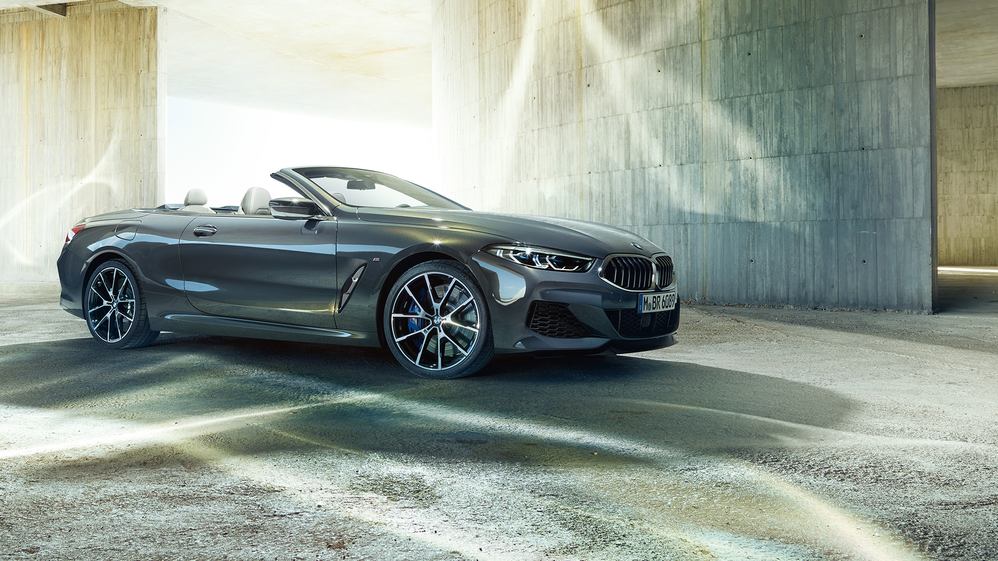 BMW M850i xDrive Cabrio, vista de tres cuartos del lateral F91. BMW M850i xDrive Cabrio en Dravitgrau metalizado con la capota abierta, vista de tres cuartos del lateral, parado delante de un edificio de hormigón F91.