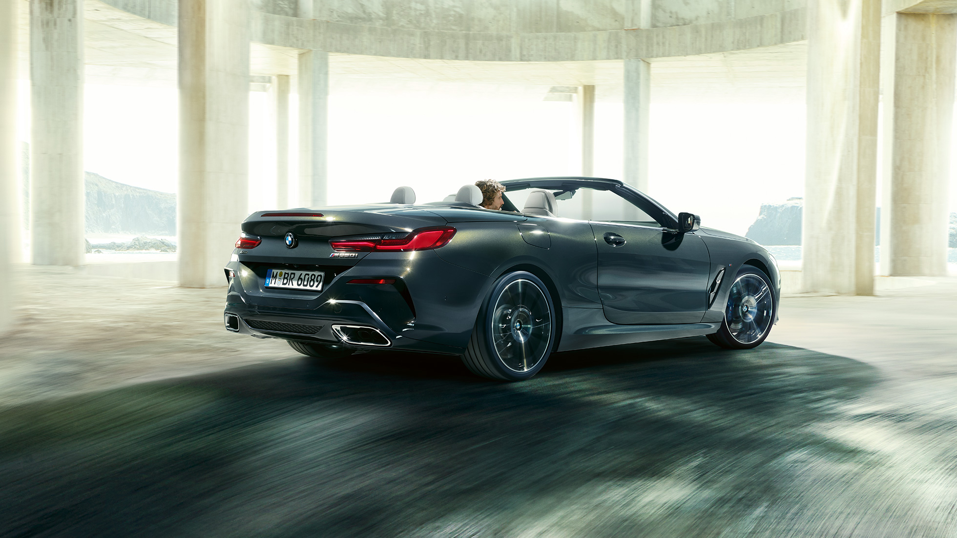 BMW M850i xDrive Cabrio, vista de tres cuartos de la zaga F91. BMW M850i xDrive Cabrio en Dravitgrau metalizado con la capota abierta, vista de tres cuartos de la zaga, atravesando un edificio de hormigón F91.