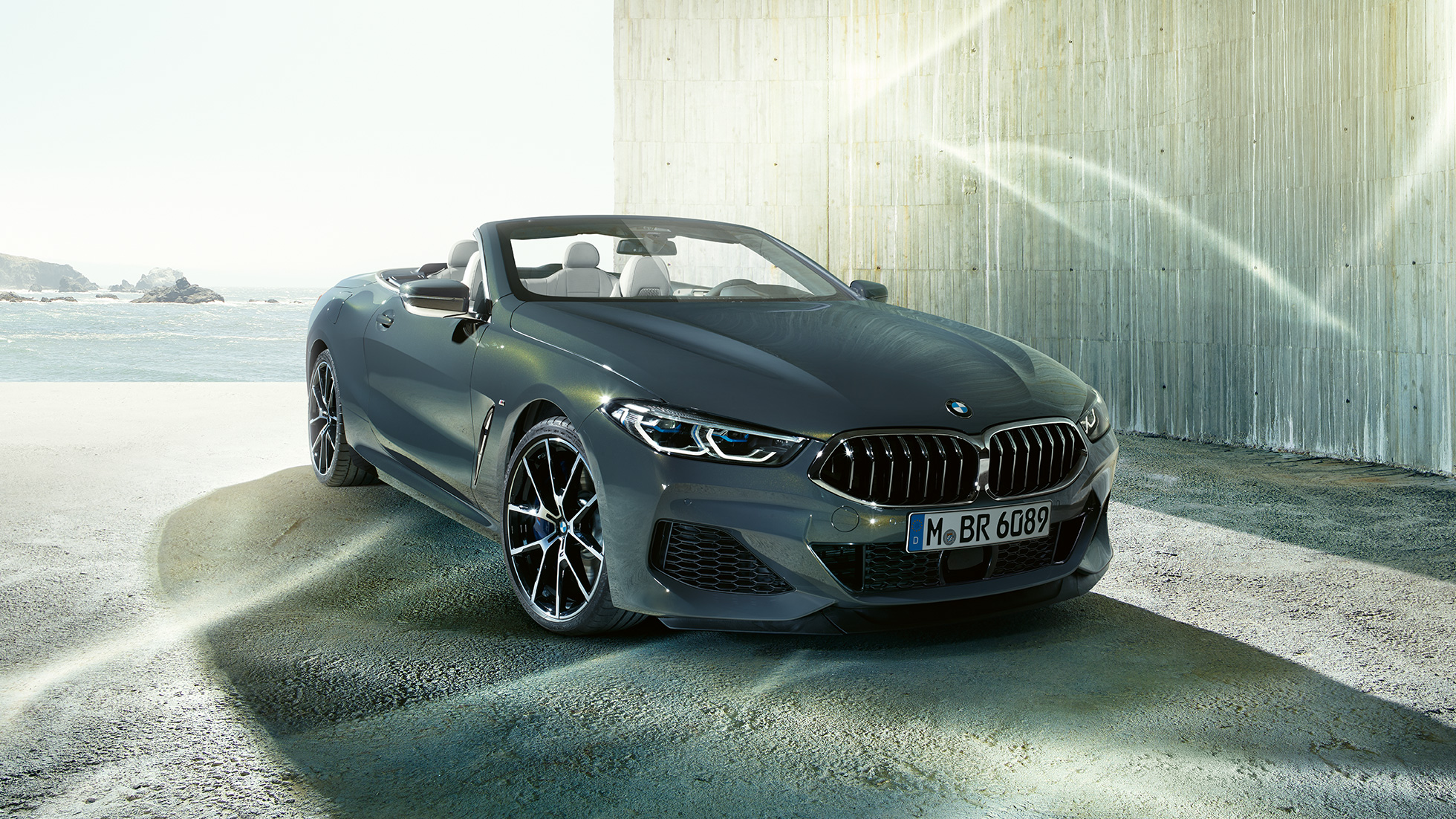 BMW M850i xDrive Cabrio, vista de tres cuartos del frontal F91. BMW M850i xDrive Cabrio en Dravitgrau metalizado con la capota abierta, vista de tres cuartos del frontal, parado delante de la costa F91.