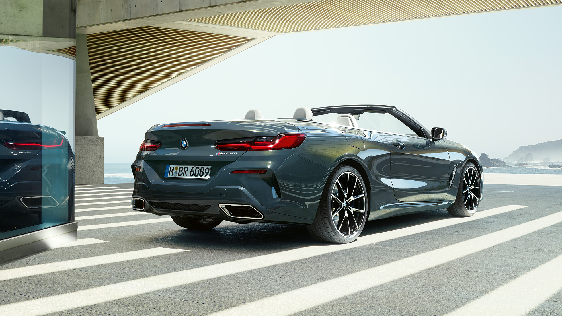 BMW M850i xDrive Cabrio, vista de tres cuartos de la zaga F91. BMW M850i xDrive Cabrio en Dravitgrau metalizado con la capota abierta, vista de tres cuartos de la zaga, aparcamiento con vista al mar F91.