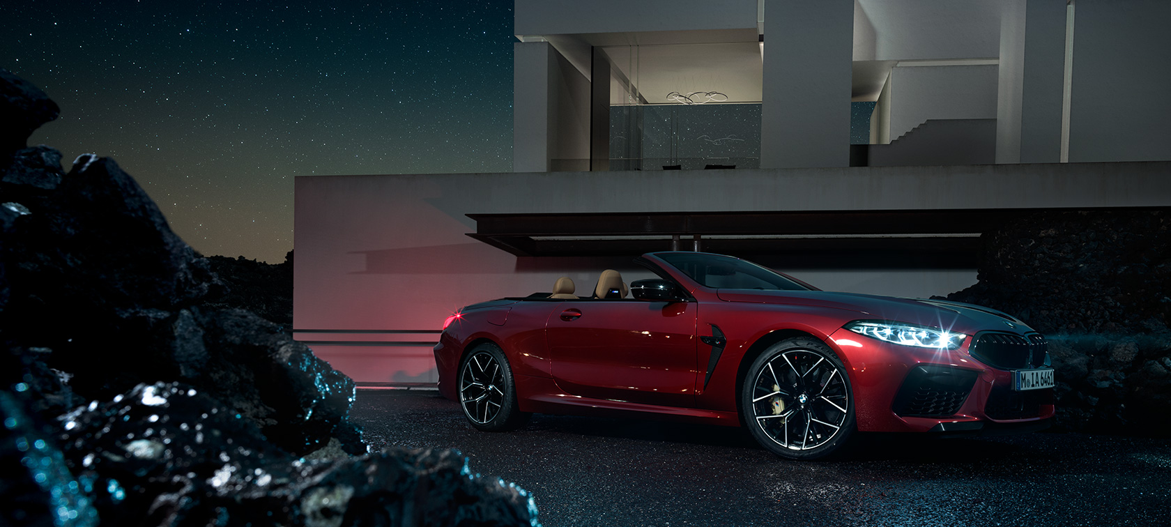 BMW M8 Competition Cabrio, vista de tres cuartos del lateral. BMW M8 Competition Cabrio en Motegi Rot metalizado, vista de tres cuartos del lateral, parado delante de una casa de campo, de noche.