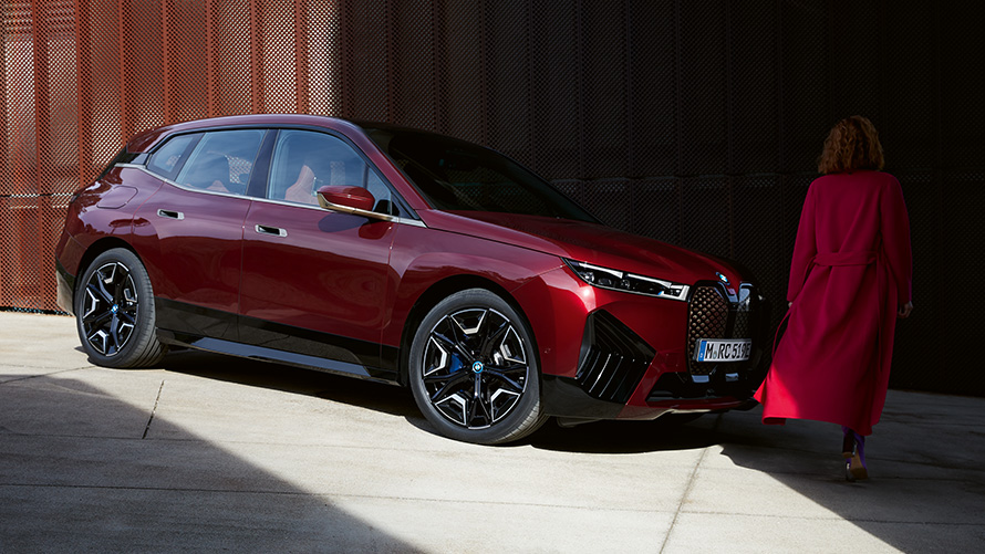 Mujer delante del BMW iX i20 2021, vista de tres cuartos del frontal Mujer vestida con abrigo rojo delante del BMW iX i20 2021, vista de tres cuartos del frontal
