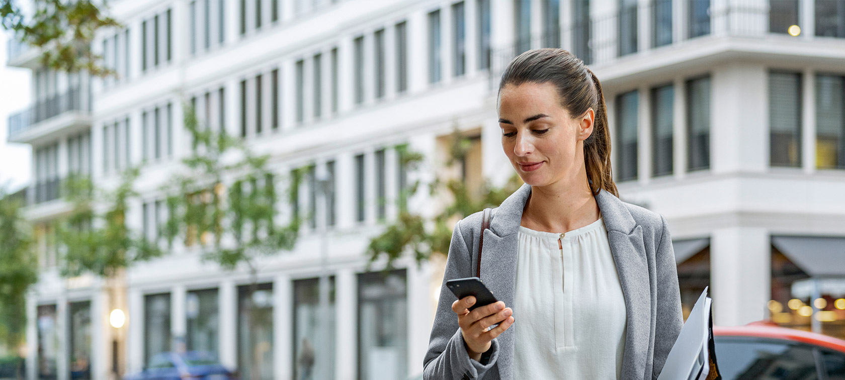Una mujer, situada delante de un edificio, mira su smartphone Remote Software Upgrade, mujer delante de un edificio que mira su smartphone