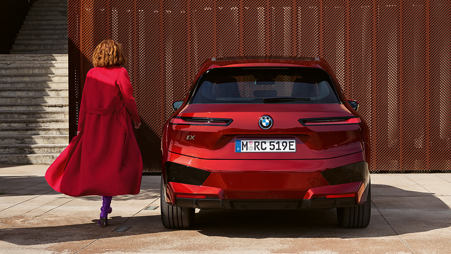 Mujer con abrigo rojo junto a un BMW iX (i20), cierre del portón trasero Cierre del portón trasero, mujer con abrigo rojo junto a un BMW iX (i20) delante de un edificio