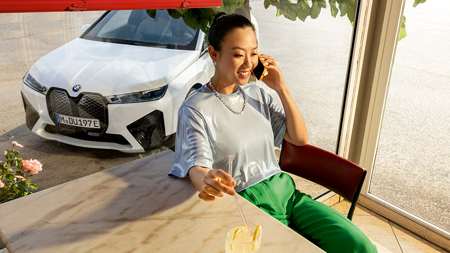 Requisitos Proactive Care Proactive Care, Revisión y mantenimiento Requisitos de Proactive Care Proactive Care, Revisión y mantenimiento, mujer sentada en la mesa con smartphone en la mano y con un vehículo BMW al fondo